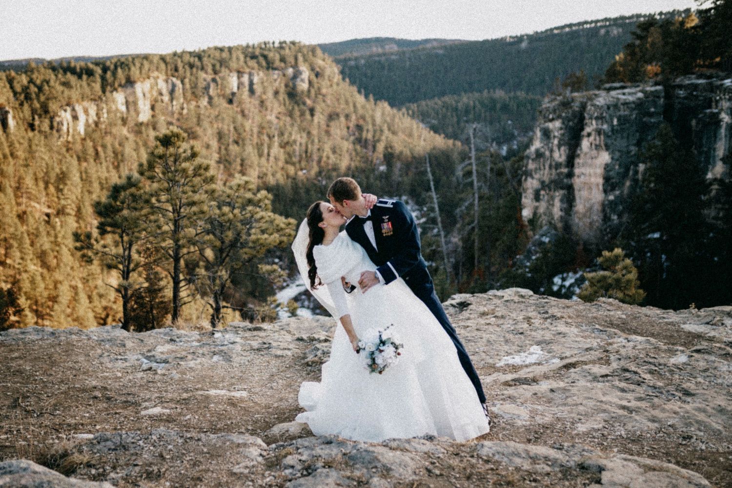 Bride and groom share romantic dip kiss on rocky mountain overlook with forested canyon backdrop.