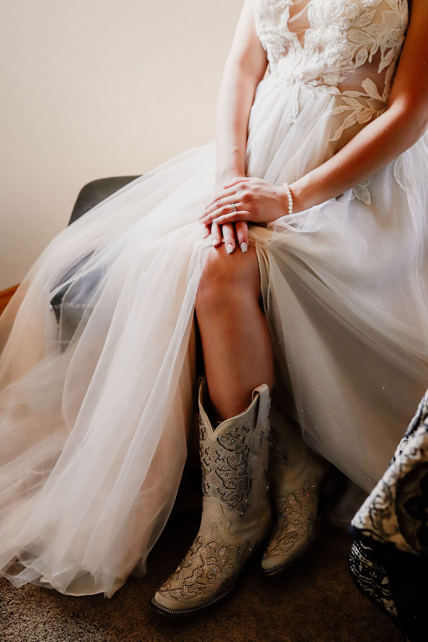Bride in lace wedding gown and white cowboy boots sits elegantly, showcasing her diamond ring and pearl bracelet.
