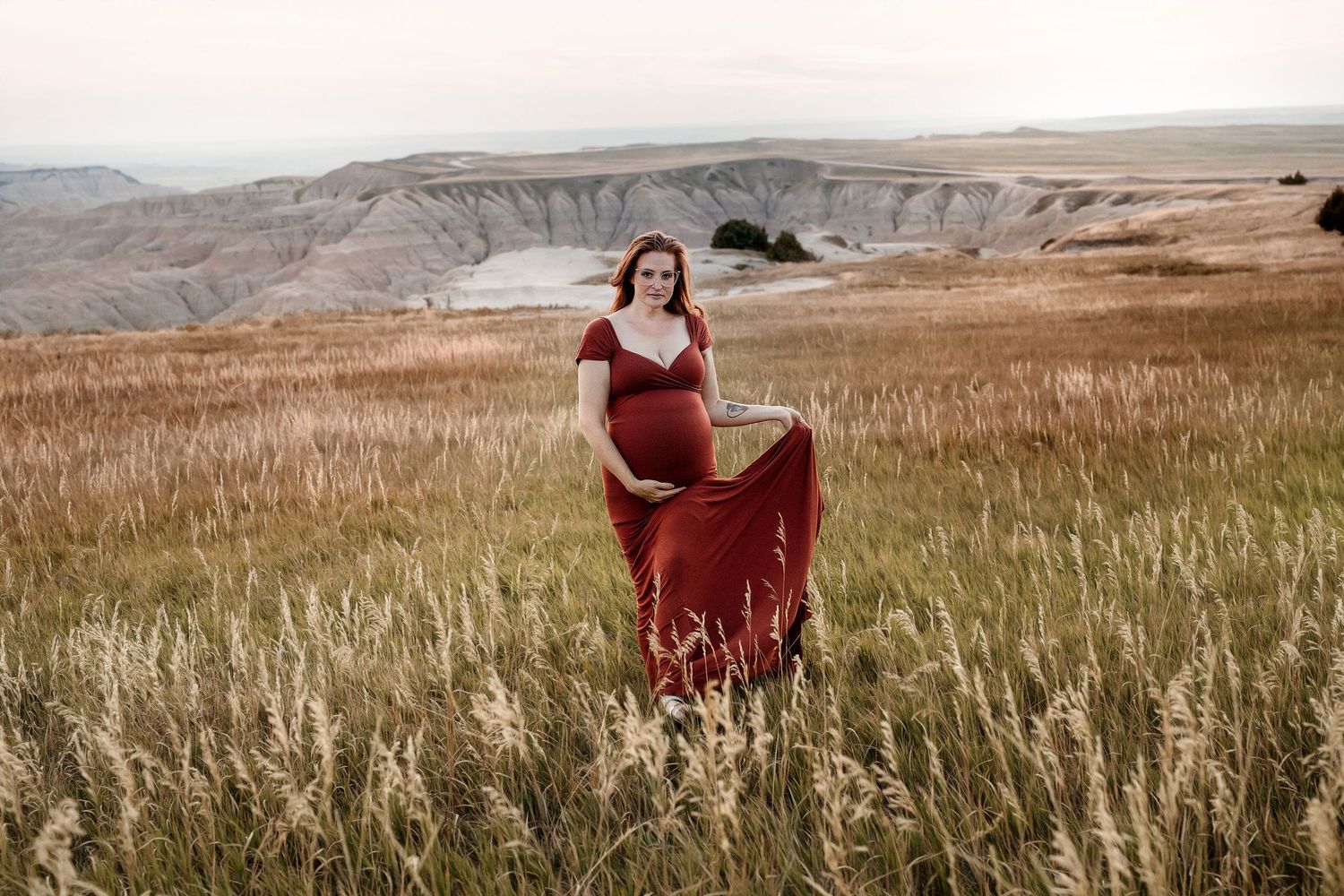 Pregnant woman in red dress walks through golden grass with badlands cliffs behind her.