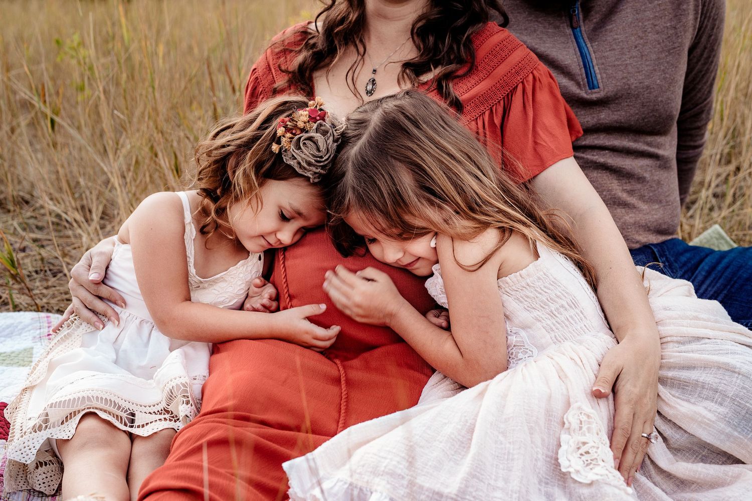 Couple with two young daughters cuddle together on blanket in golden grass field.