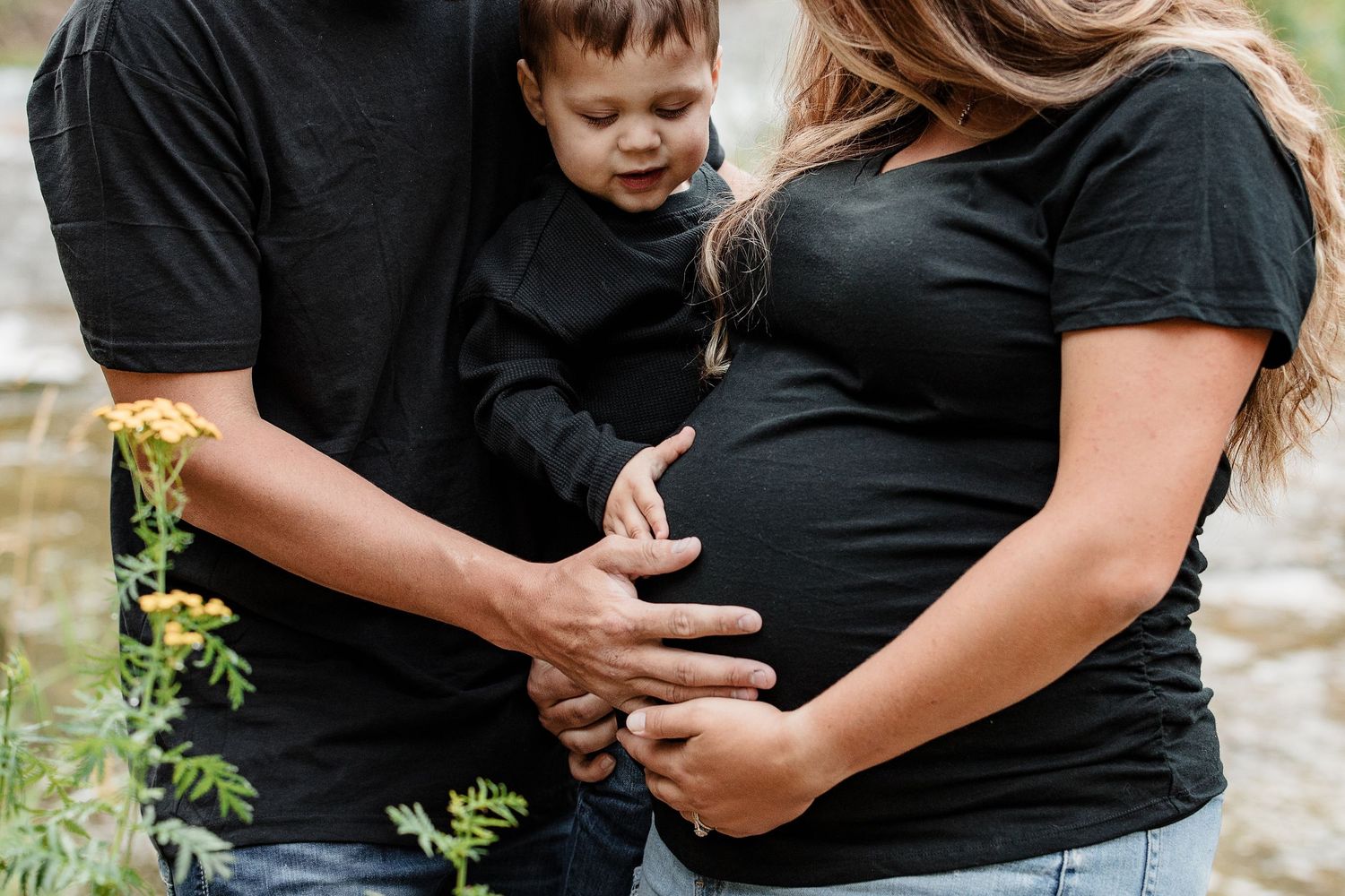 Close-up of pregnant woman in black top with partner and toddler touching baby bump near wildflowers.