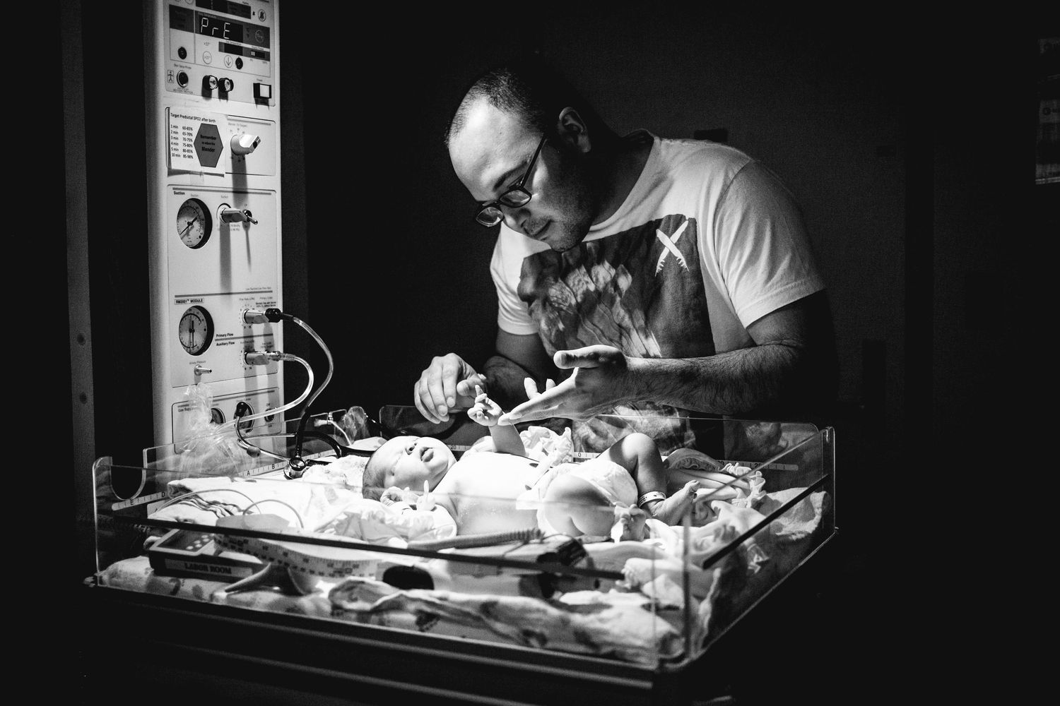 Father leaning over his newborn baby in a hospital warming bassinet in black and white.