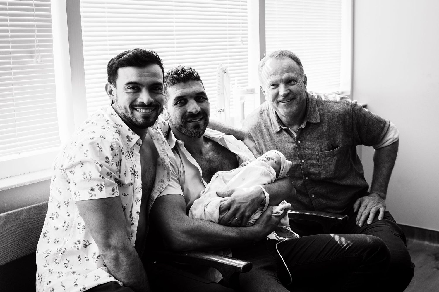 Three men smile together holding newborn baby in hospital room, black and white family birth photo.
