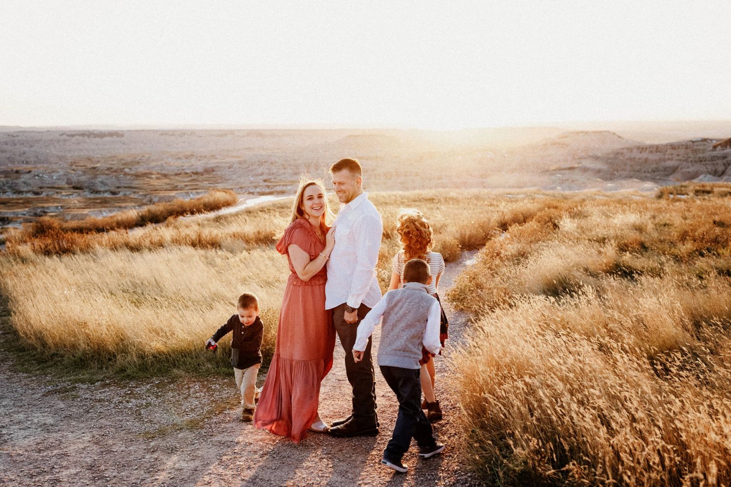 Family of five standing together on a dirt path in a golden field at sunset.