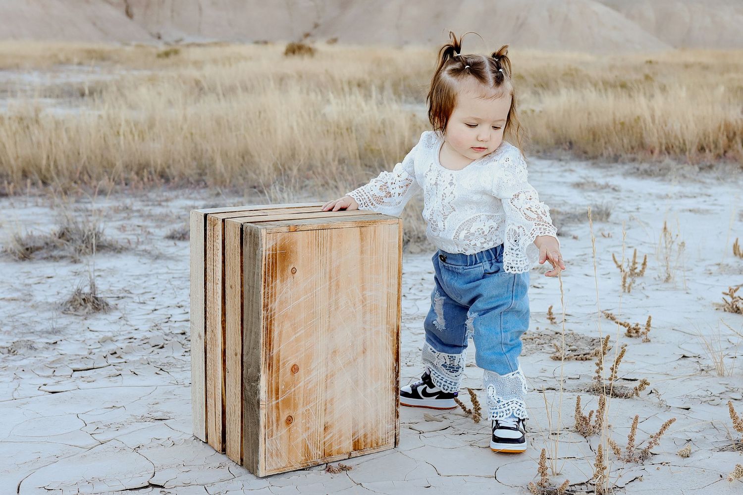 Toddler girl in lace top and jeans standing next to a rustic wooden crate on sandy desert terrain.