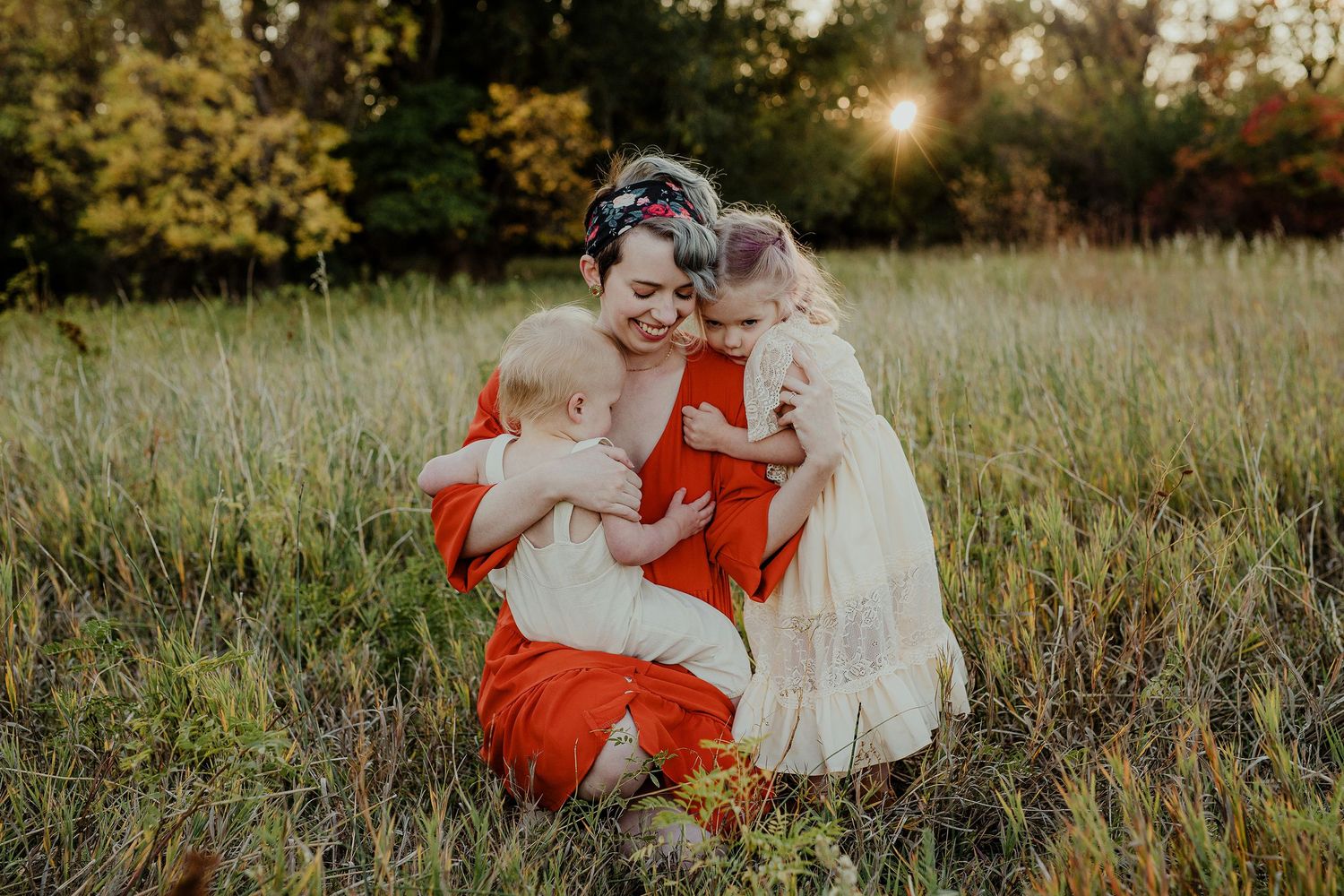 Mother in red dress hugging two young children in a sunlit meadow during golden hour.