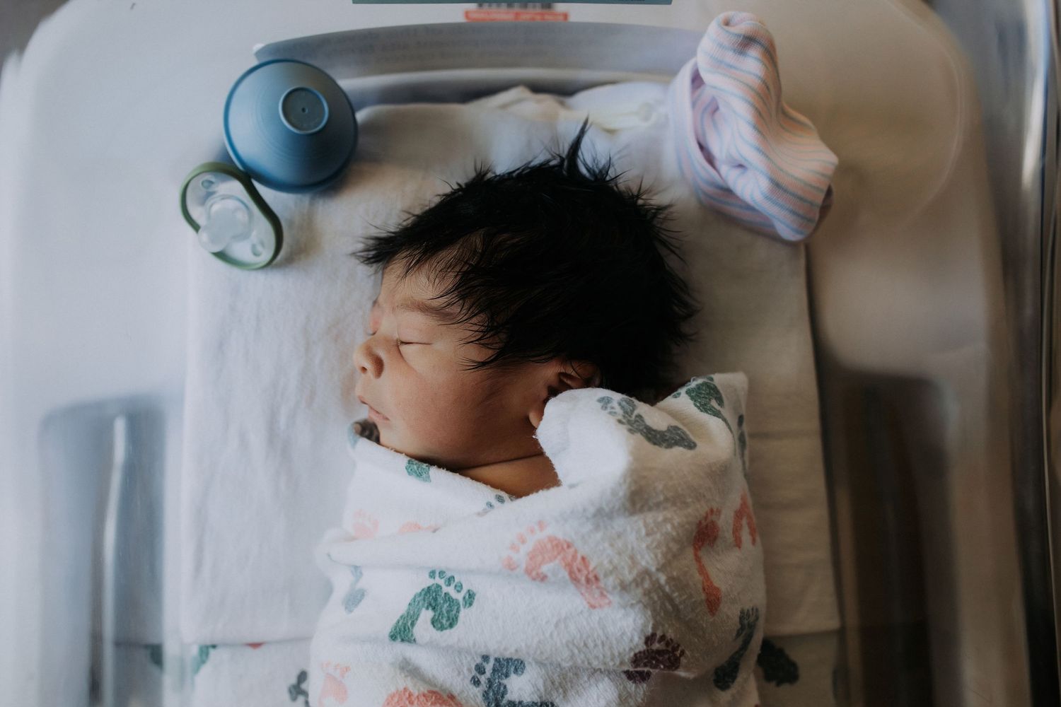 Newborn baby with dark hair swaddled in a colorful blanket sleeping in a hospital bassinet.