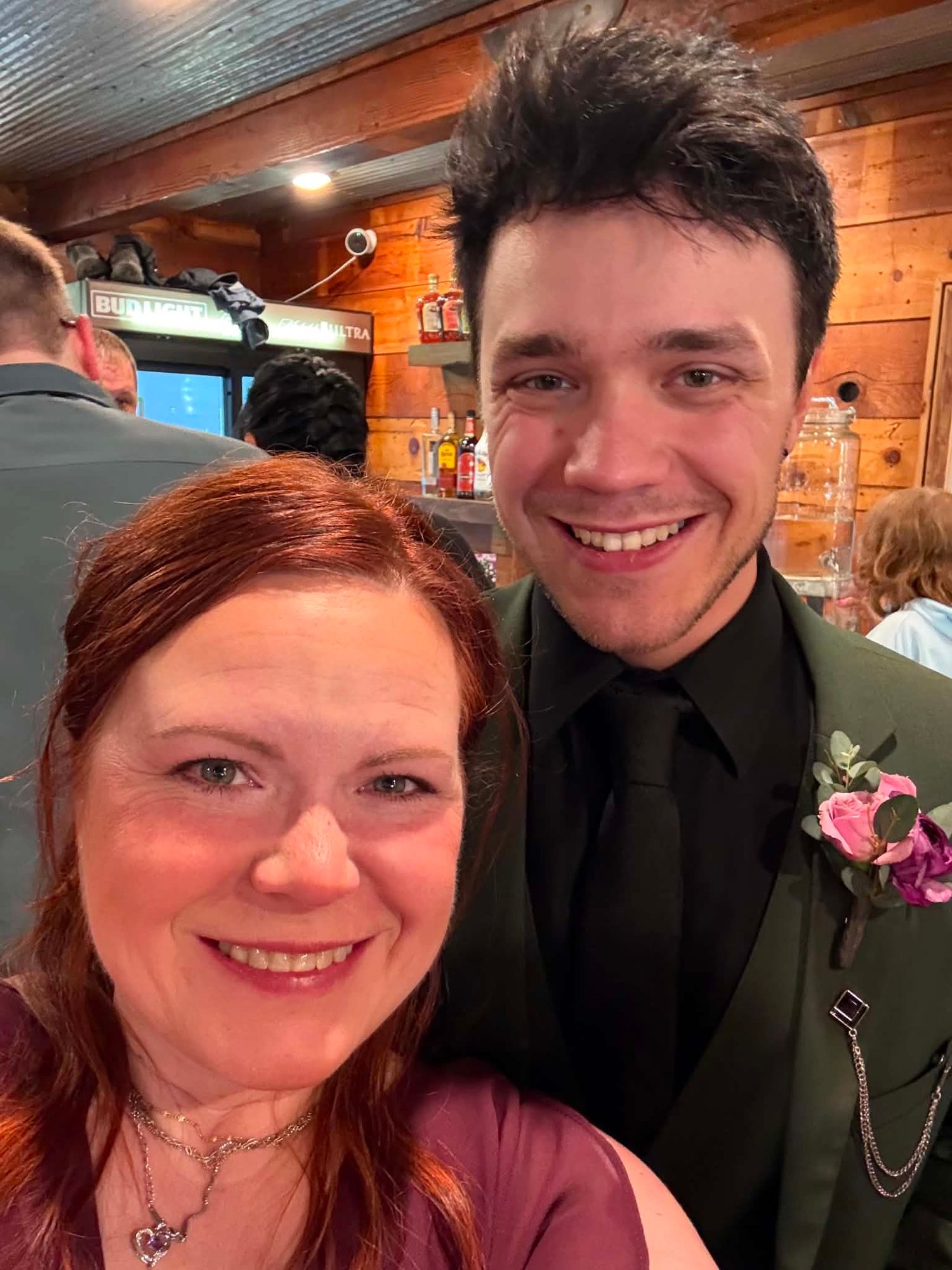 Two people smiling at a formal event, woman in burgundy dress and young man in green suit with boutonniere.