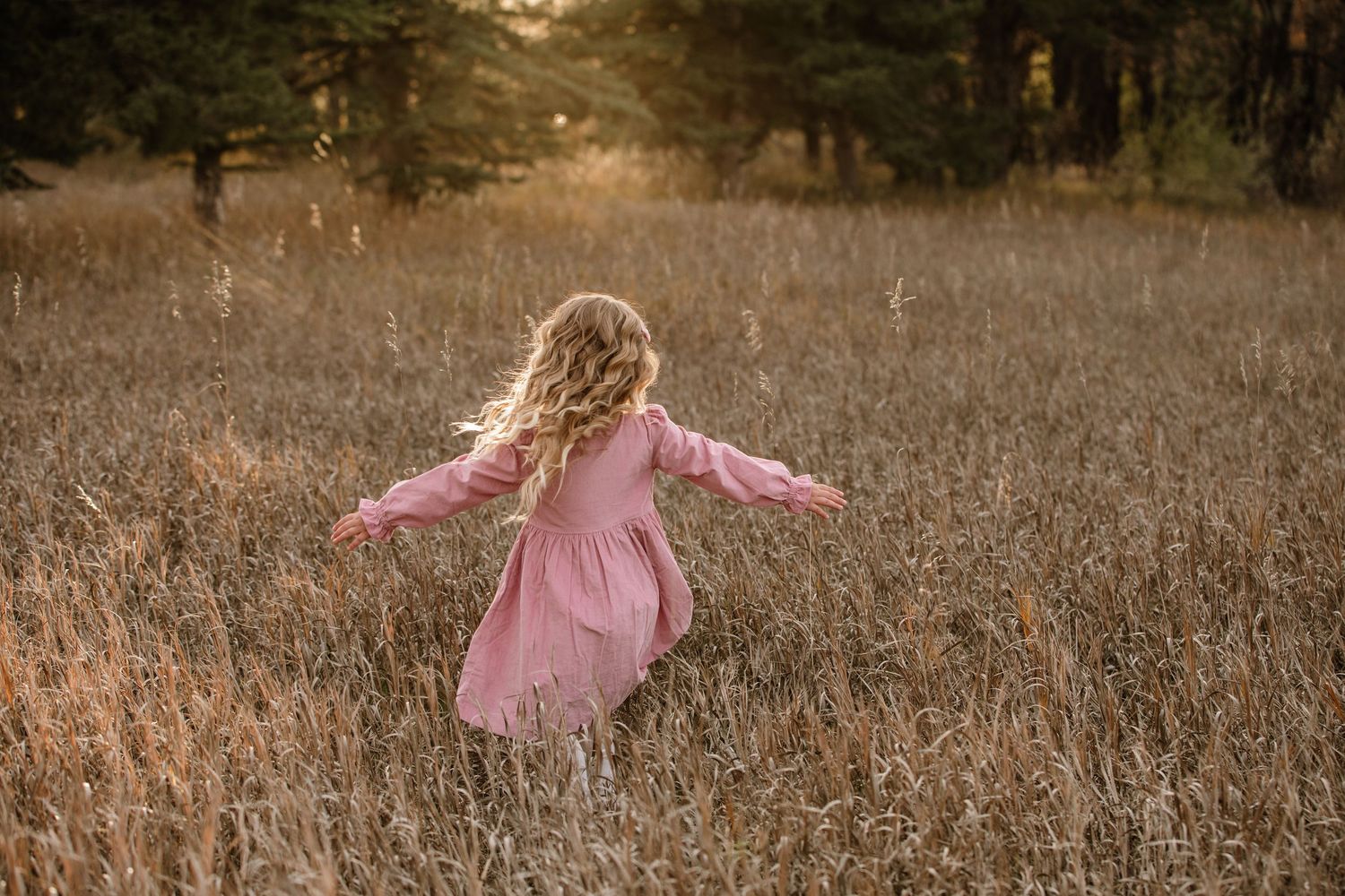 A young girl in a pink dress runs through a golden field at sunset with her curly hair flowing behind her.