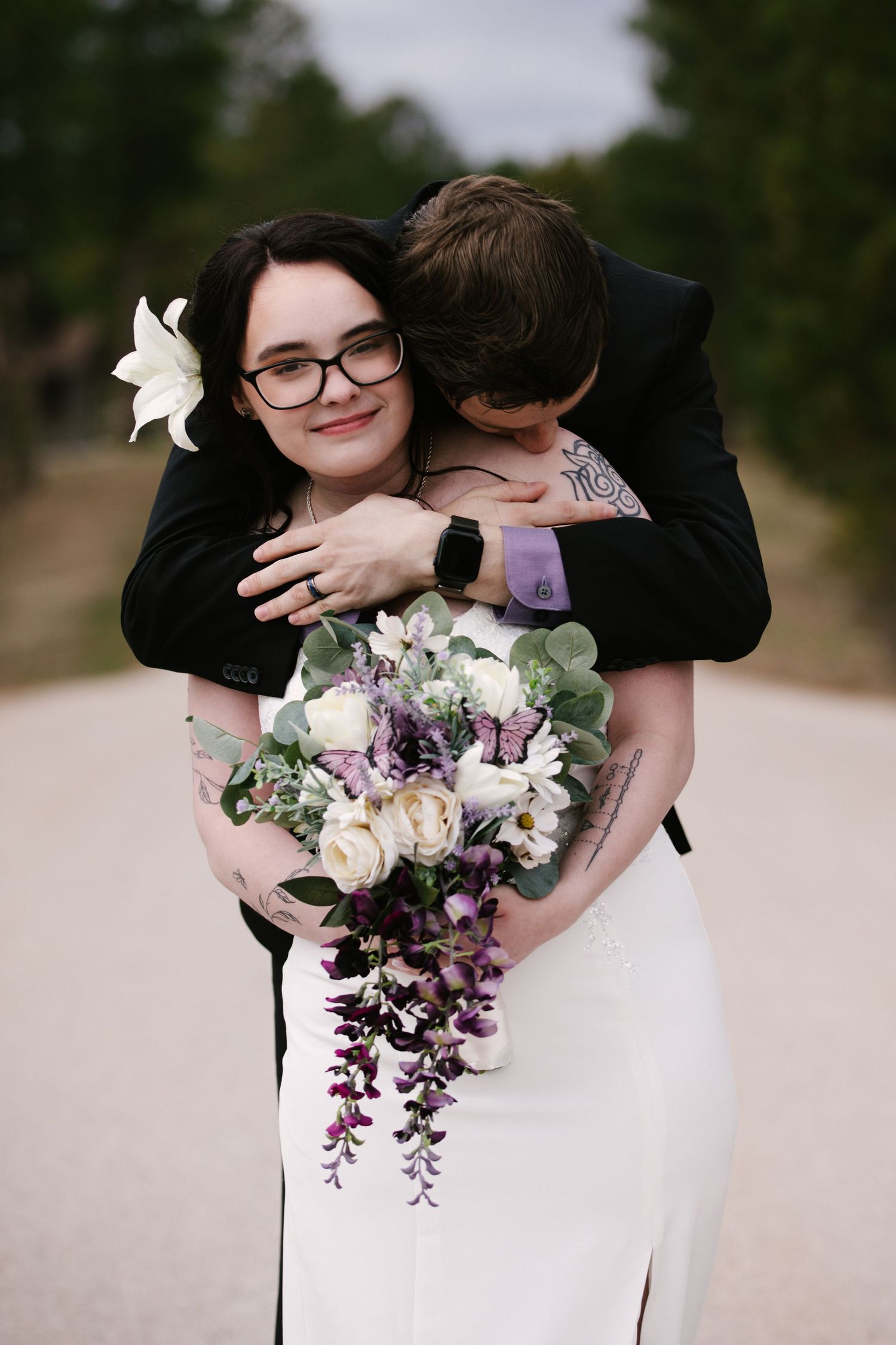 Bride in white dress holding purple and white bouquet, embraced by groom in black suit on a scenic road.