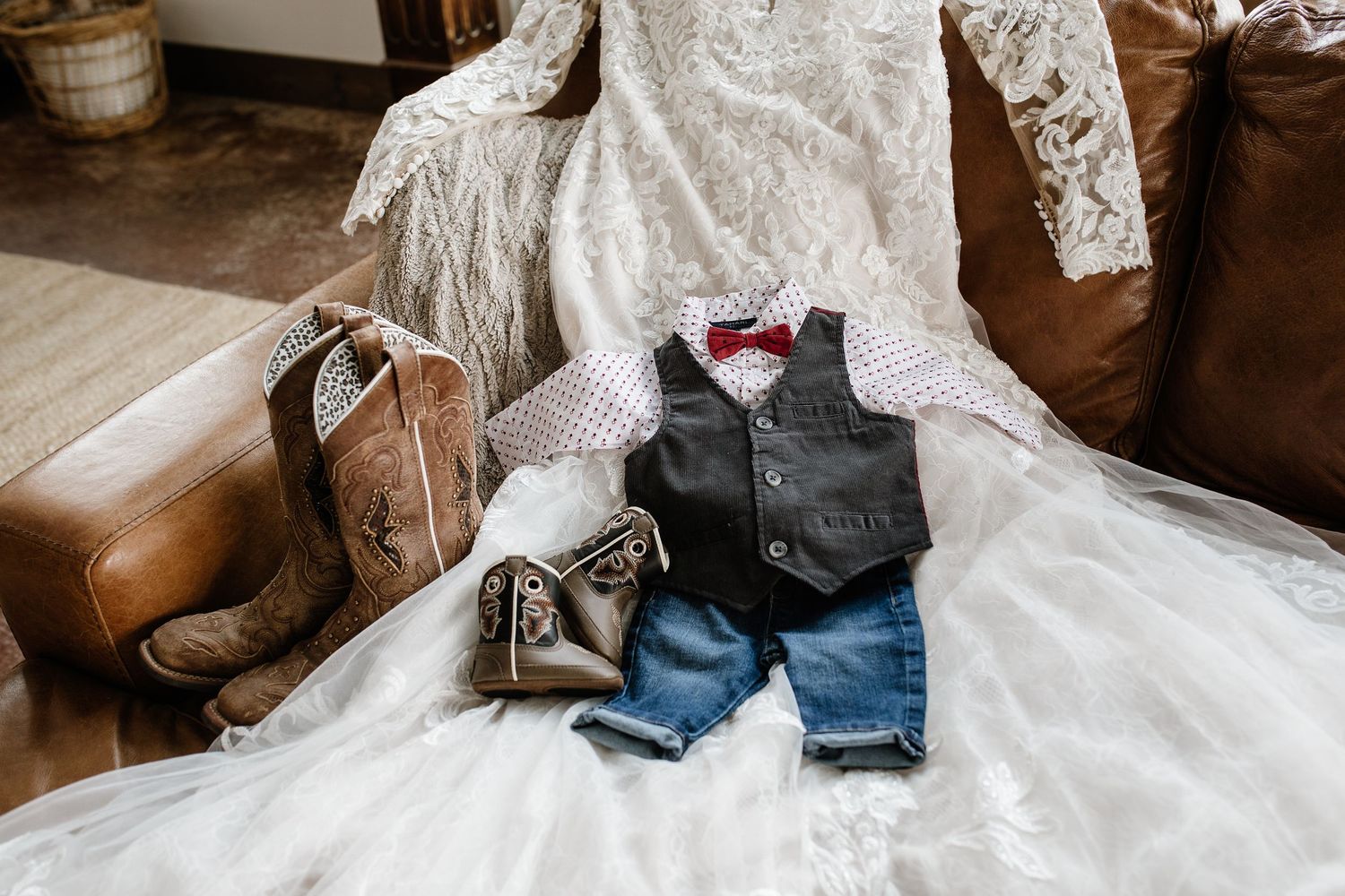 Wedding flat lay with lace bridal gown, cowboy boots, and ring bearer outfit featuring vest, bow tie, and jeans.