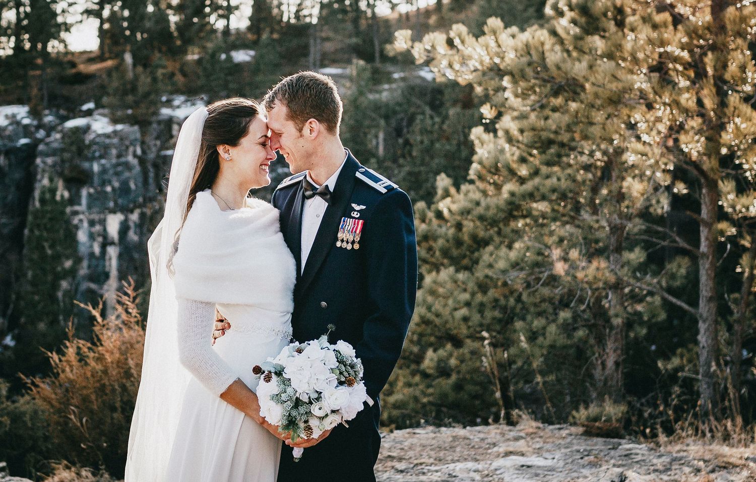 Military groom in dress uniform embraces bride in white gown holding bouquet outdoors at scenic rocky mountain wedding.