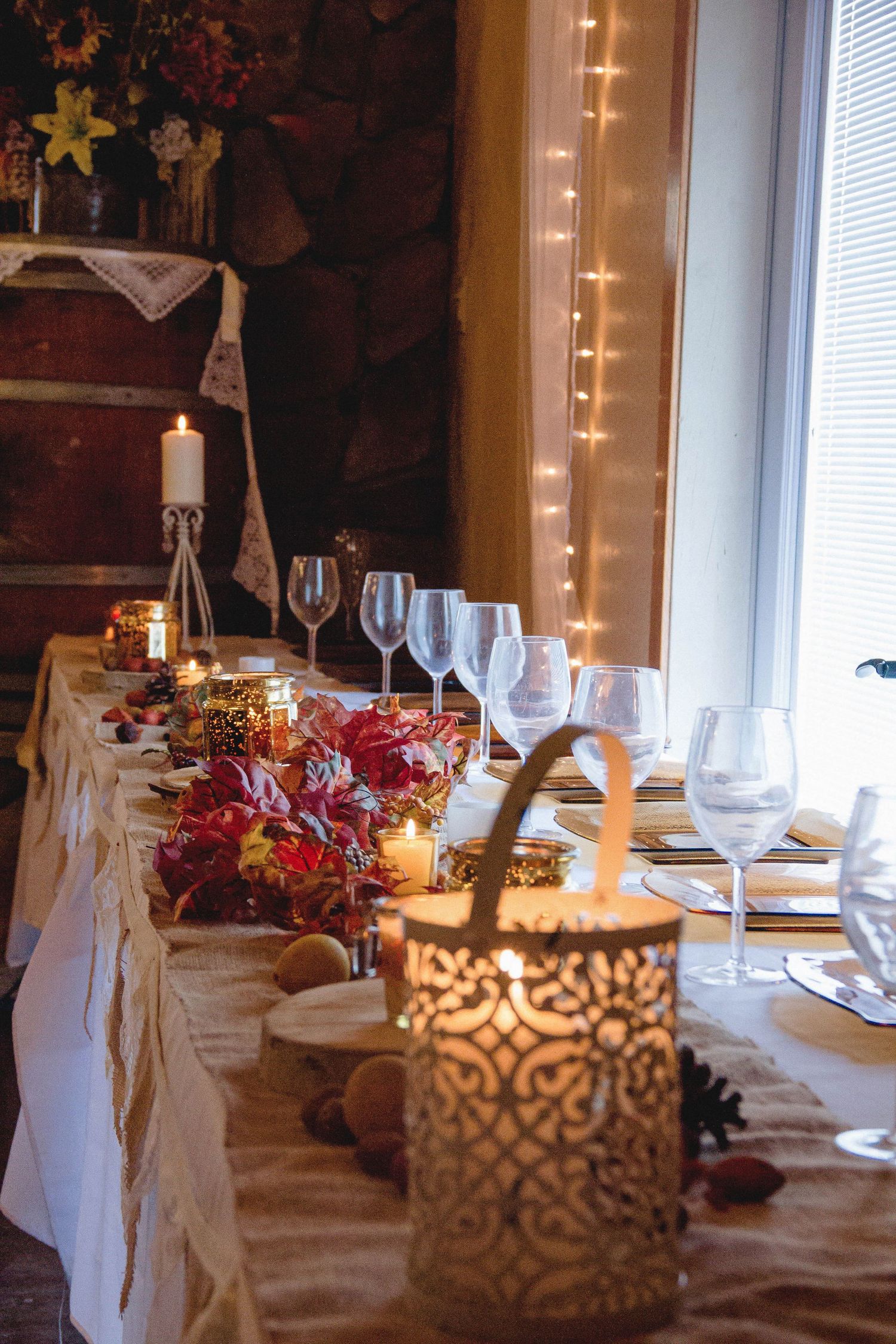 Elegant table setting with wine glasses, floral centerpieces, candles, and fairy lights in a cozy rustic dining room.
