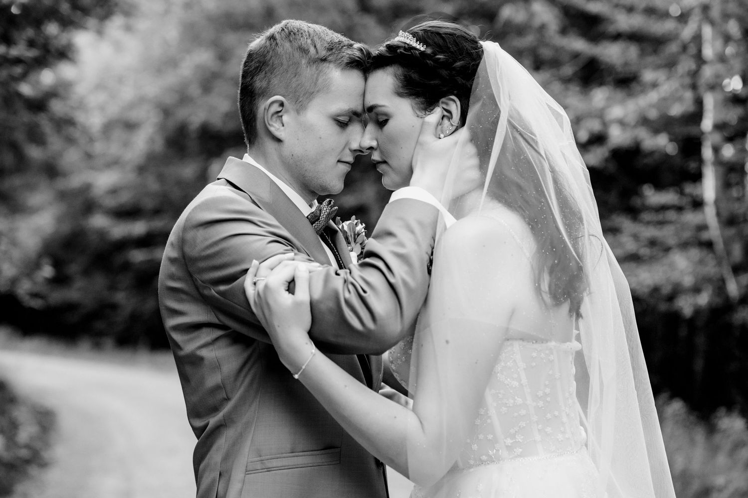 Black and white wedding photo of bride and groom sharing an intimate moment, foreheads touching, surrounded by trees.