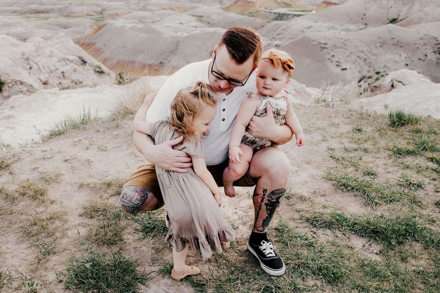 Father with glasses and tattoos crouching to hug two young daughters outdoors in badlands setting.