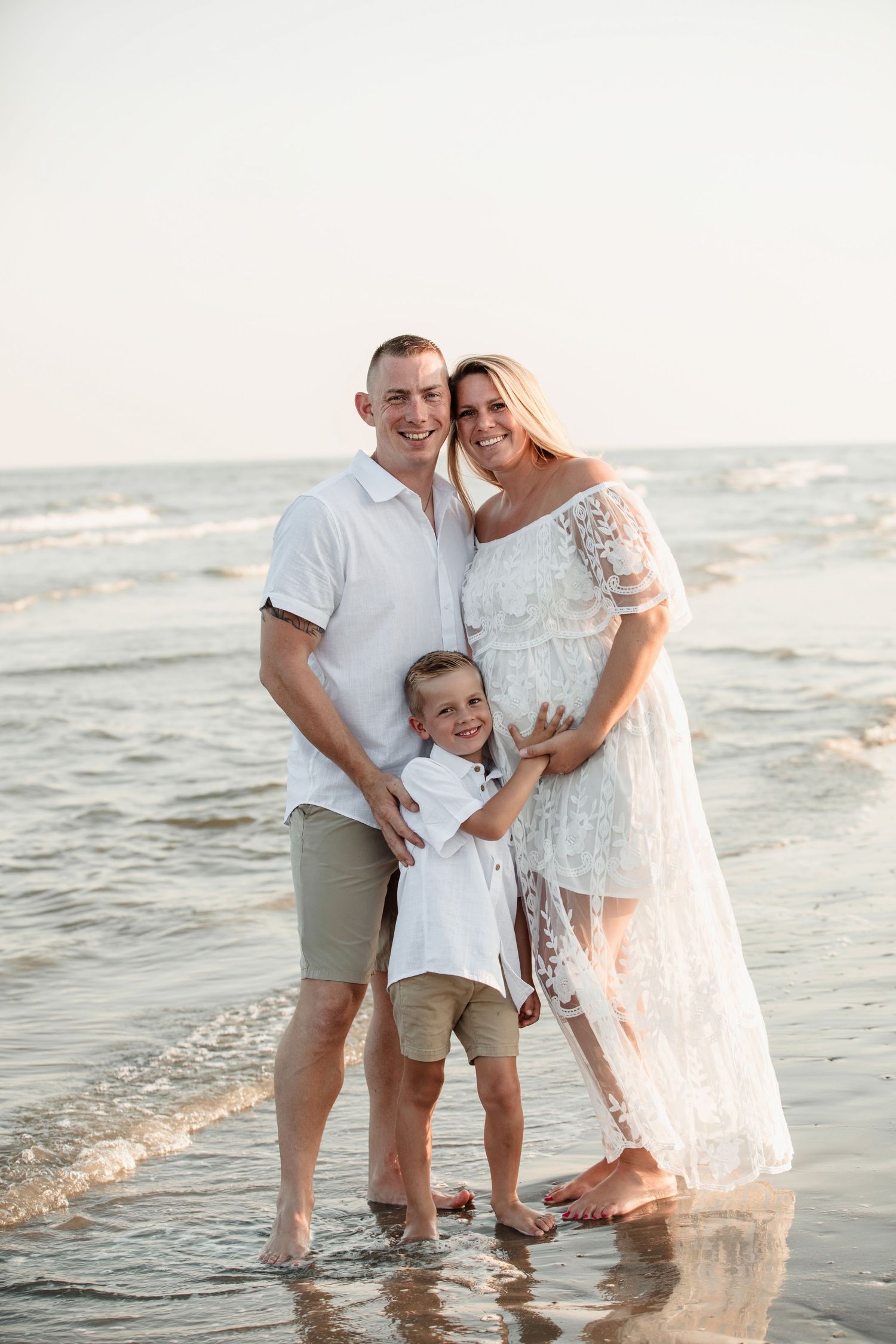Smiling family with pregnant woman in white lace dress posing with husband and son on beach.