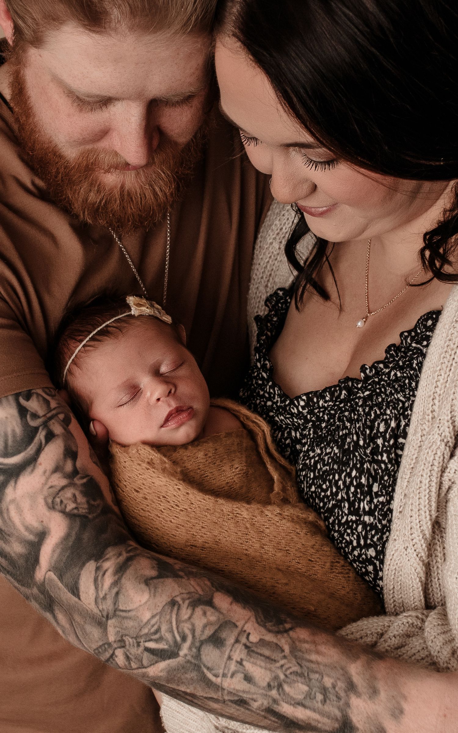 Tattooed parents lovingly gaze at their sleeping newborn wrapped in a tan swaddle blanket.