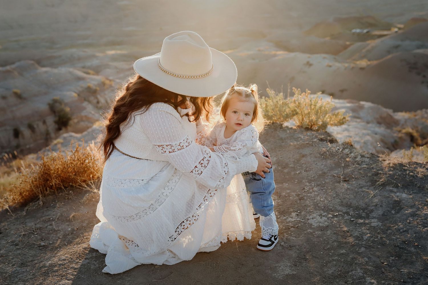 Mother in white boho dress and hat crouching with baby girl in sunny desert landscape.