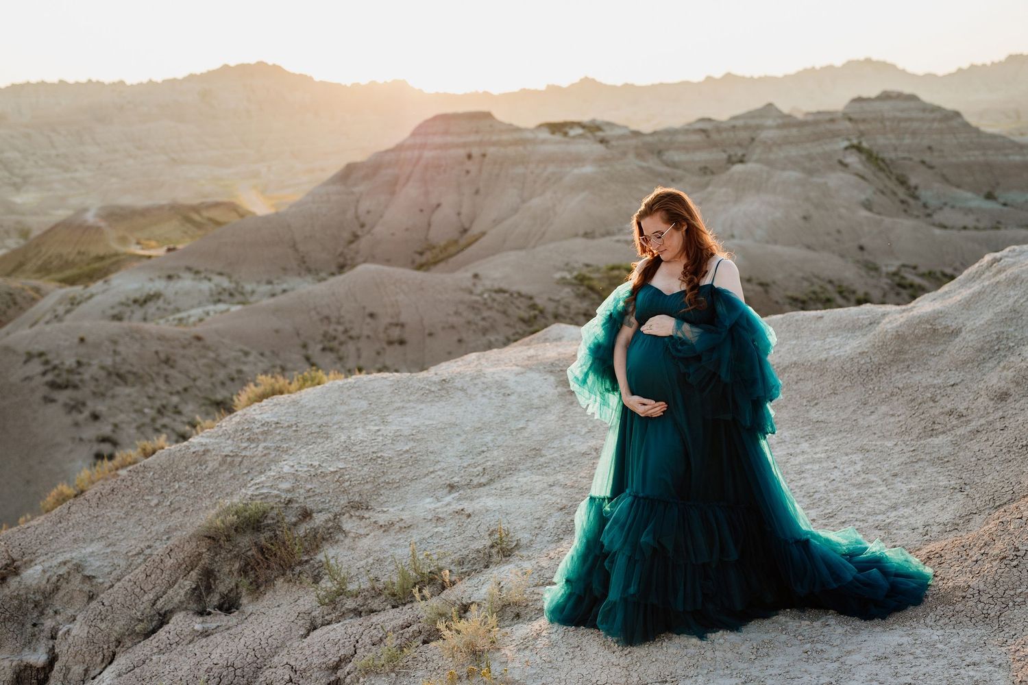 Pregnant woman in teal tulle gown cradles bump at golden hour in desert badlands.