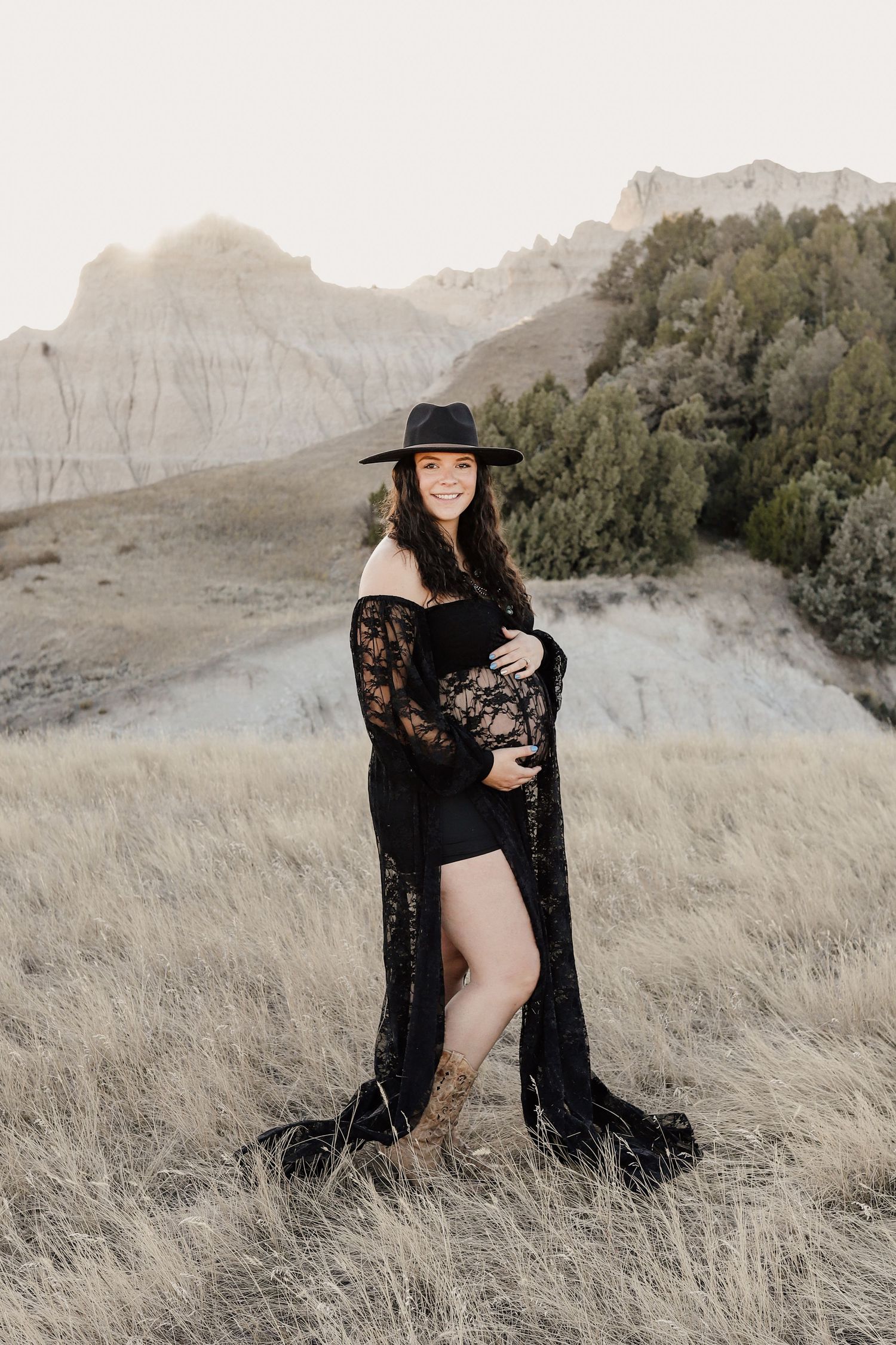 Smiling pregnant woman in black lace maternity gown and hat in badlands.