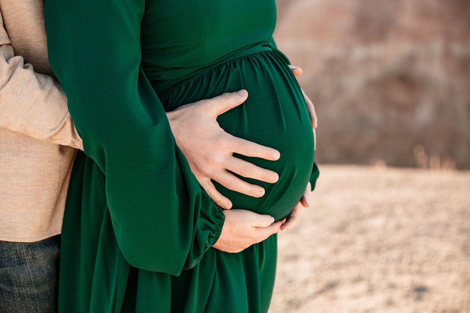 A pregnant belly in a flowing emerald green dress is tenderly cradled by hands against a soft neutral background.