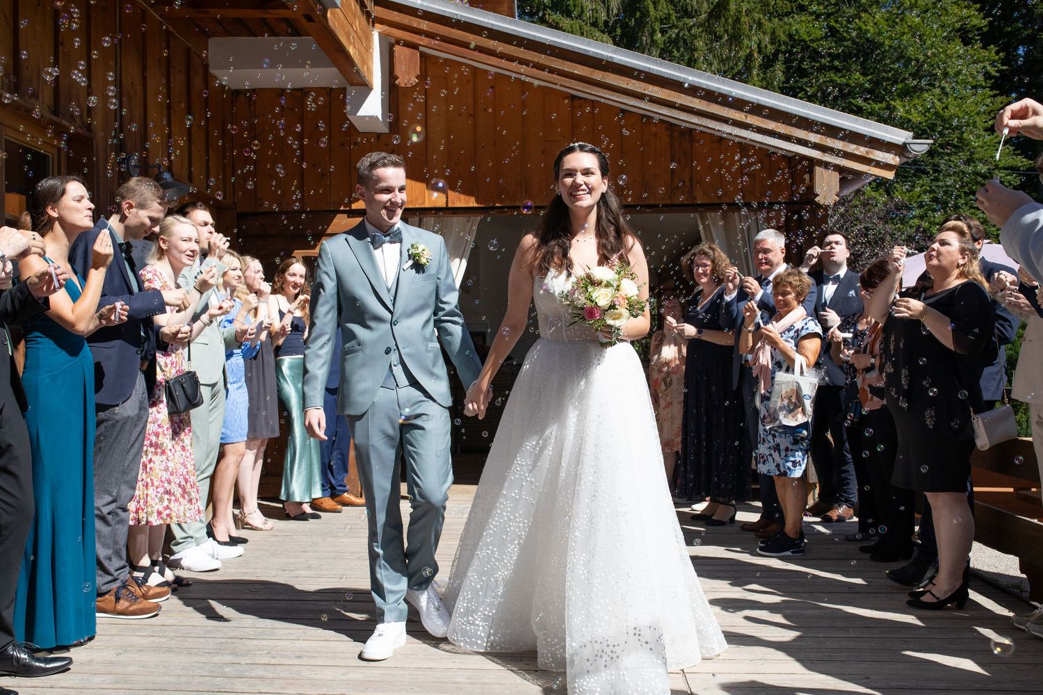 Bride and groom walk hand-in-hand through cheering wedding guests throwing confetti outdoors.