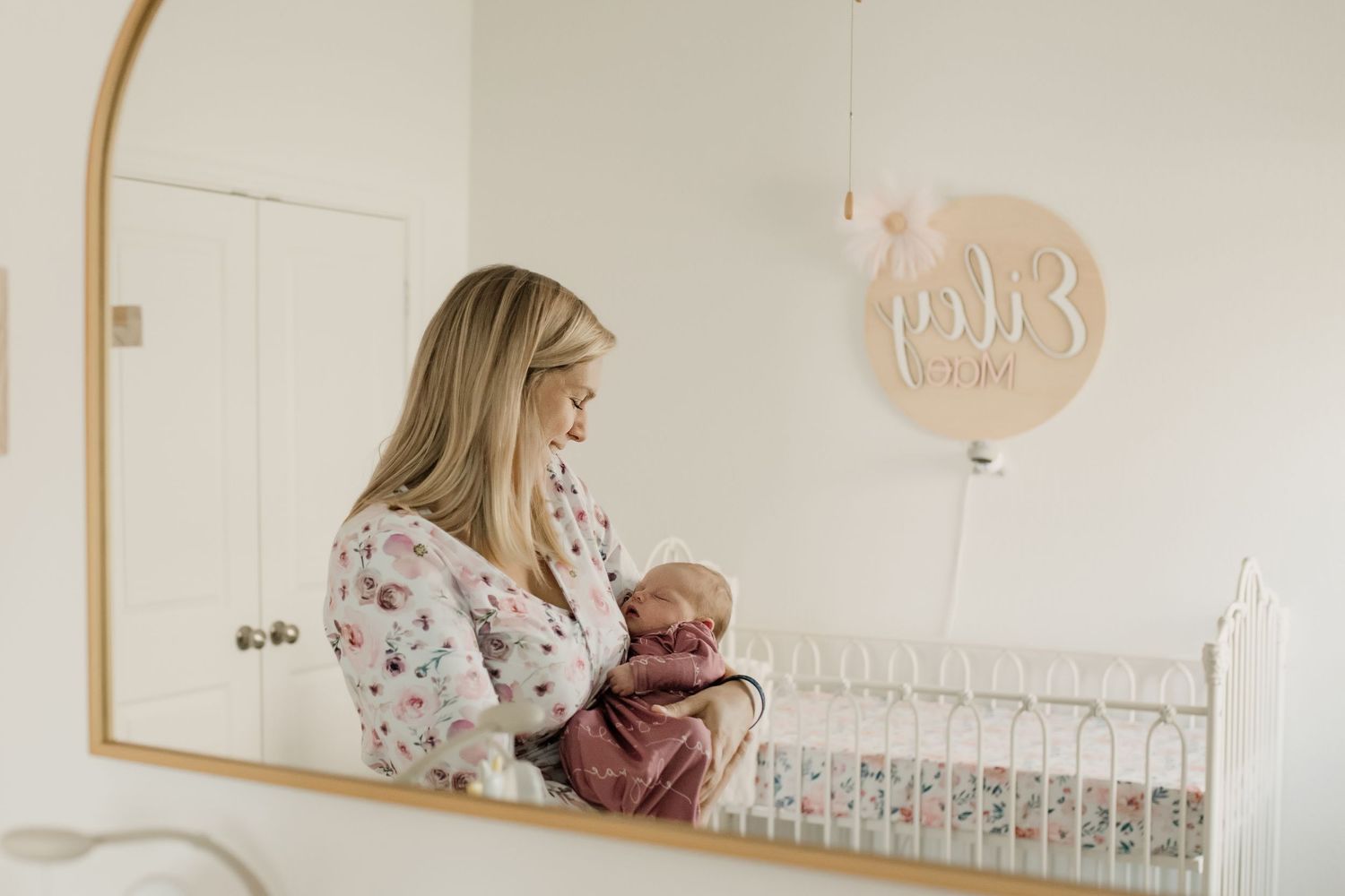 Blonde mother holding newborn baby reflected in gold arch mirror beside white iron crib in nursery.