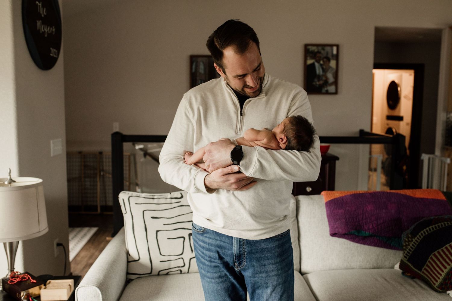 Father in white sweater cradles sleeping newborn baby while standing in a bright, modern living room.
