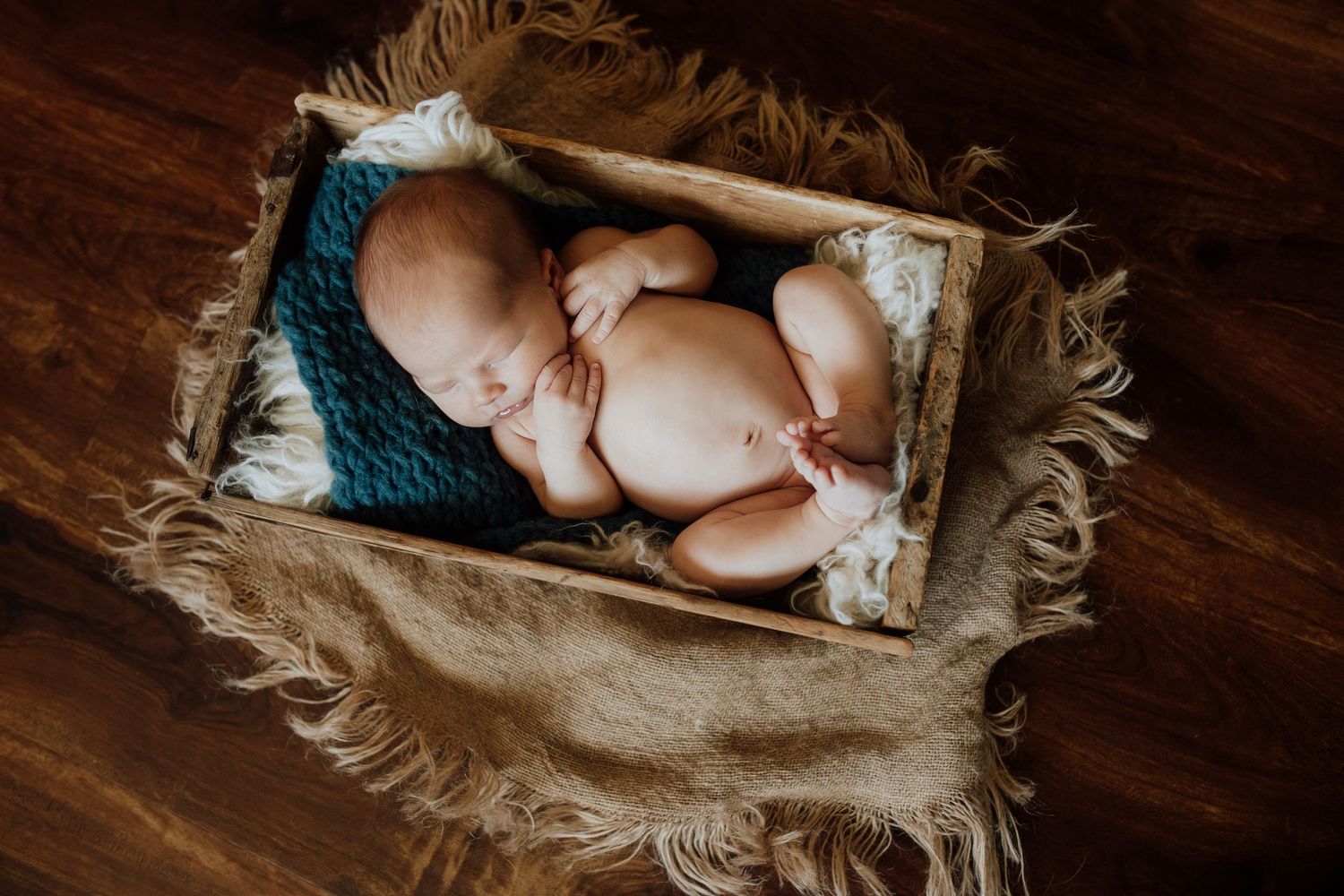 Sleeping newborn baby curled up in a rustic wooden crate with teal knit blanket on a fur rug.