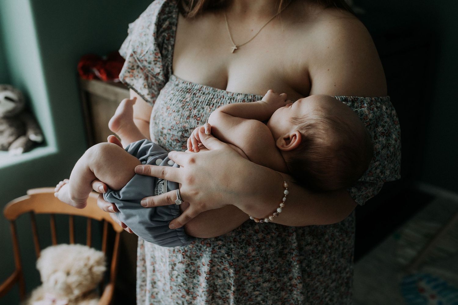 A mother in a floral dress cradles her sleeping newborn baby against her chest indoors.