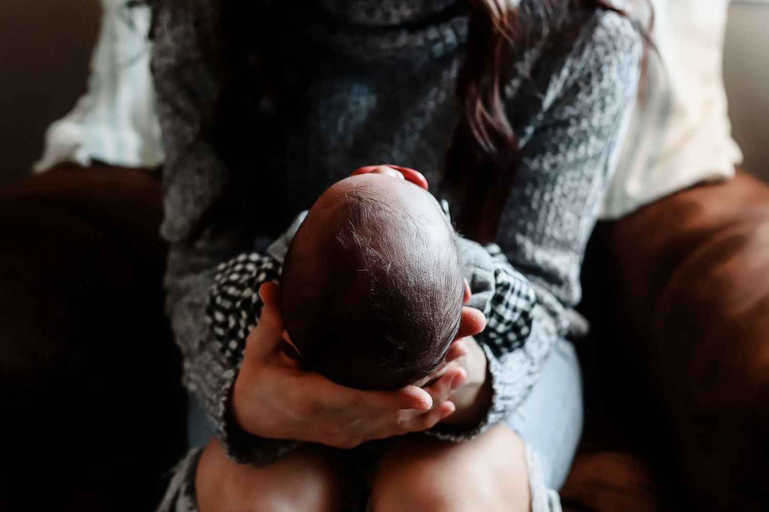 Tender overhead view of mother cradling newborns head gently in her hands while seated on brown leather chair.