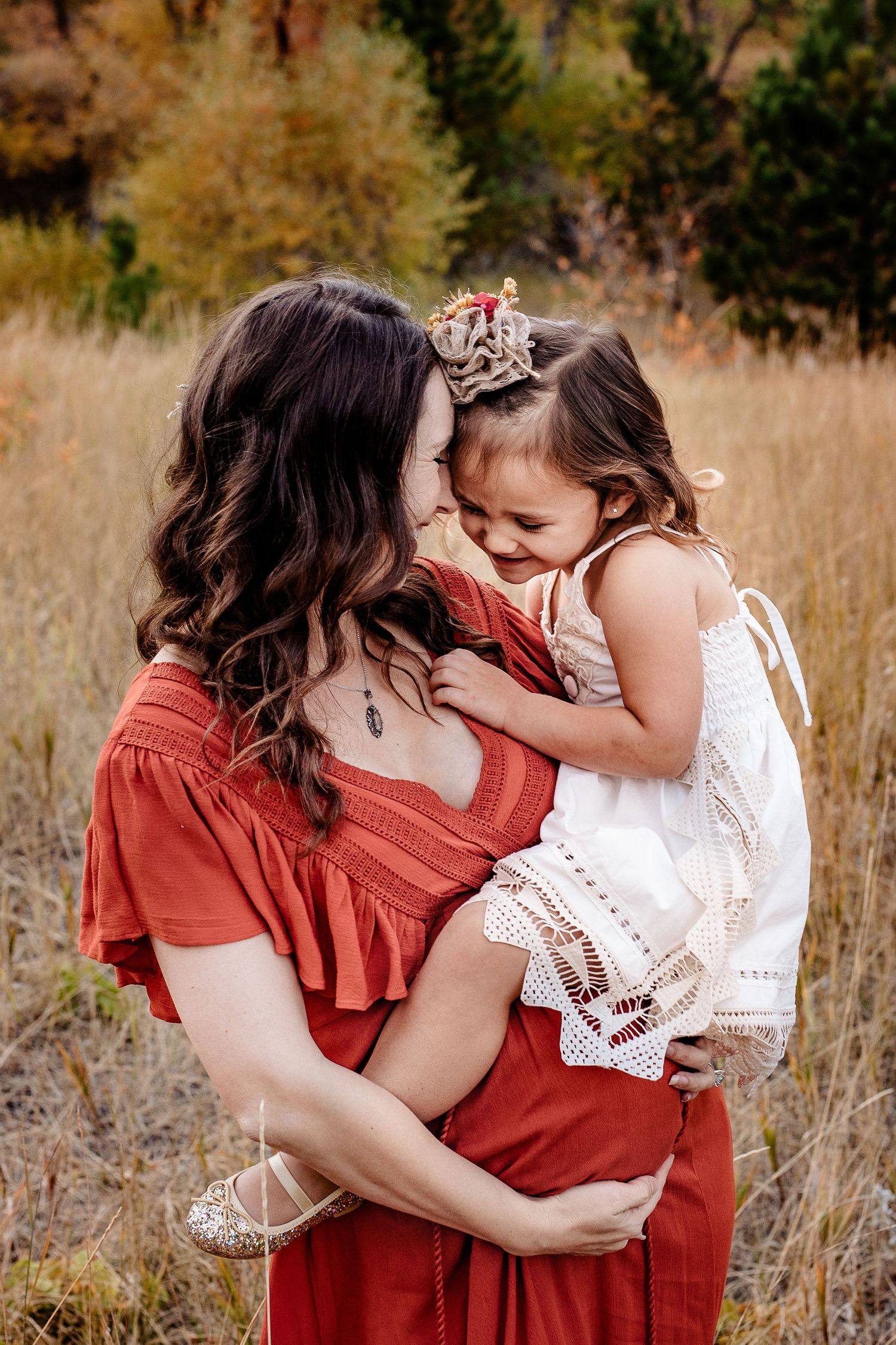 Pregnant mother in red dress holds laughing young daughter in white dress in autumn field.