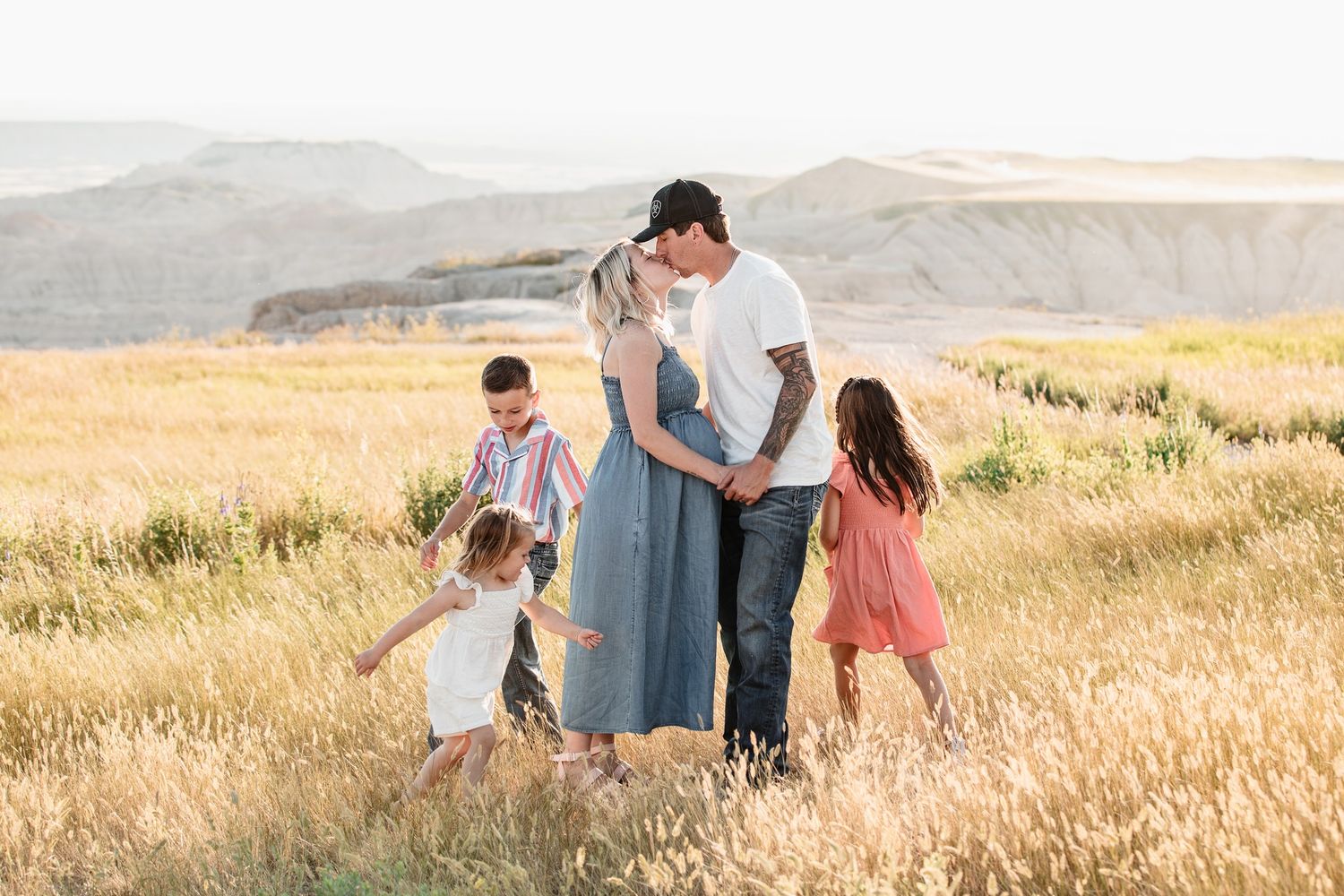 Family of five sharing a loving embrace in a sunlit golden wheat field with badlands backdrop.