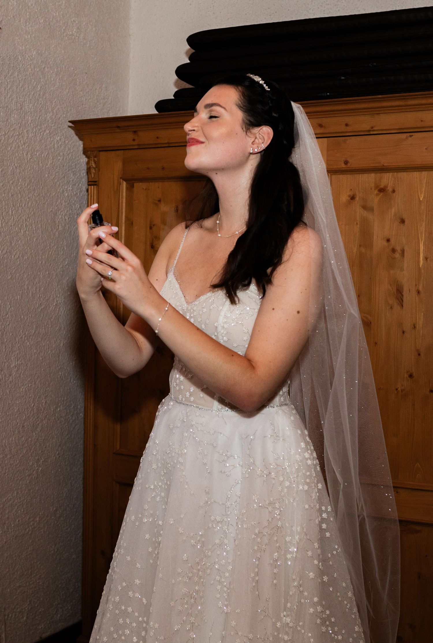 Bride in a sparkling white gown and veil, holding a small item, standing near a wooden armoire.