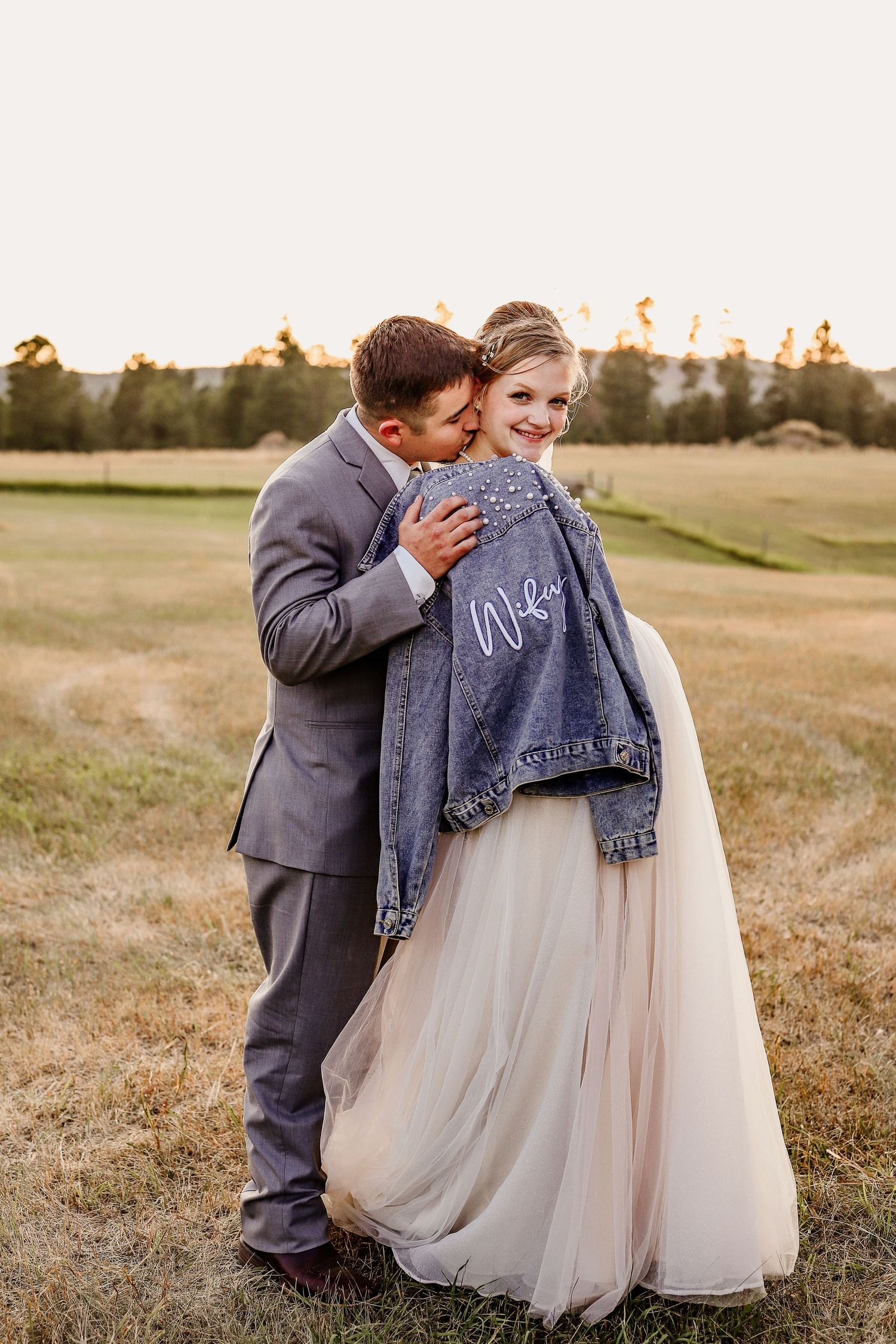 Bride in tulle skirt and denim Wife jacket embraced by groom in gray suit at golden hour outdoor wedding.