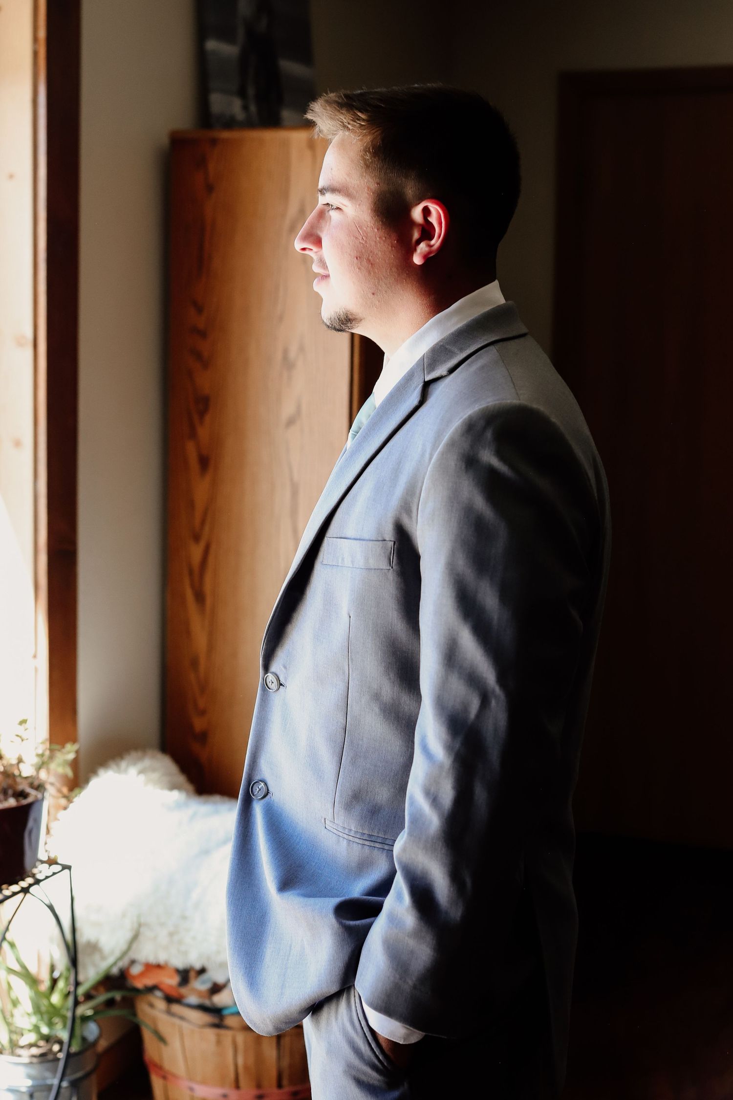 Young man in gray suit standing by window with hands in pockets, gazing thoughtfully in elegant indoor setting.