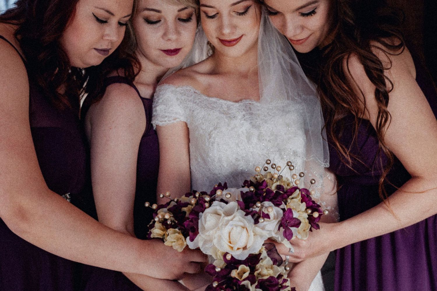 Bride in lace gown holds purple and white bouquet surrounded by bridesmaids in deep purple dresses.