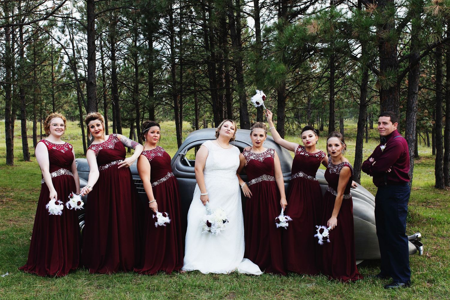 Bridal party in burgundy gowns and white bouquets posing beside a vintage car among pine trees.