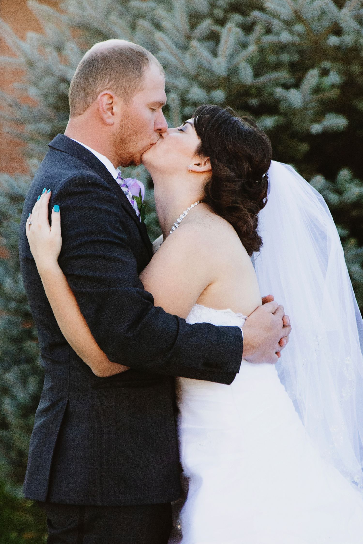 Bride and groom sharing a romantic kiss outdoors, bride in white gown with veil, groom in dark suit.