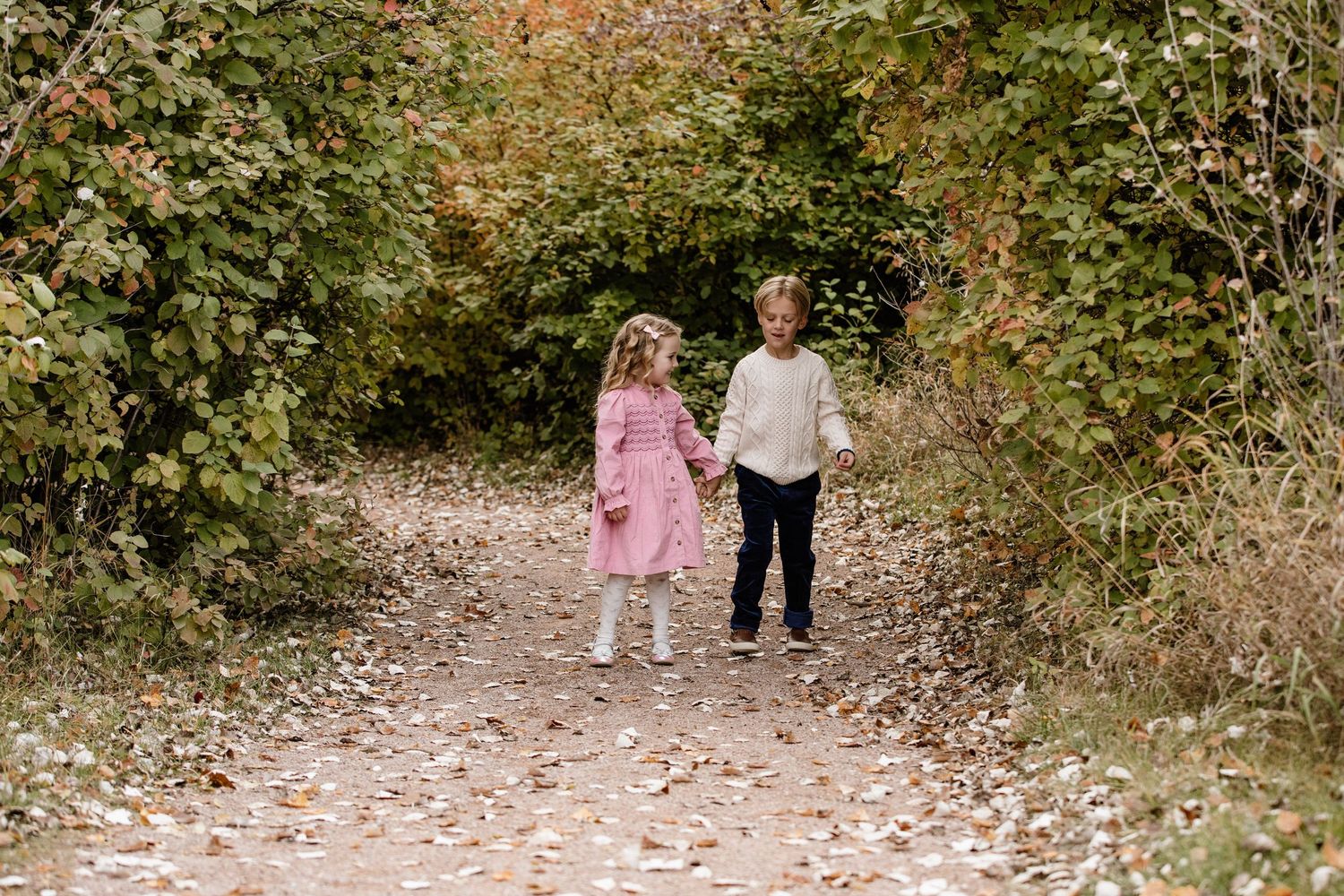 Two children holding hands on a leaf-covered autumn path surrounded by colorful fall foliage.