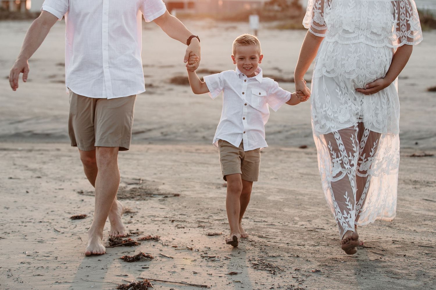 Young boy walking on beach holding hands with parents dressed in white and khaki at golden hour.