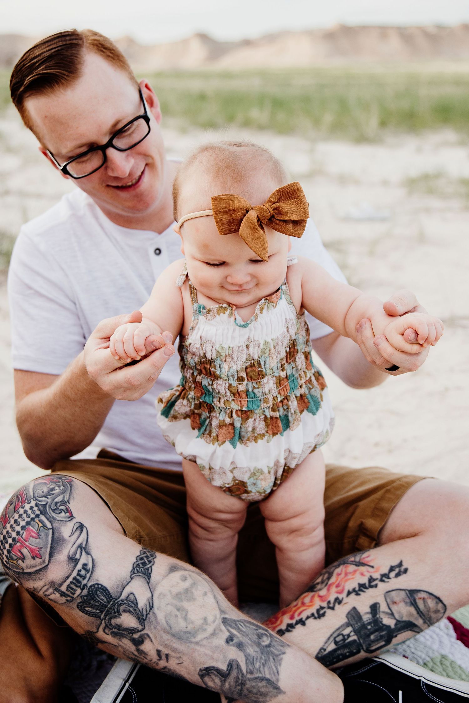 Tattooed father helping smiling baby girl in fringe romper and bow headband stand on his lap.