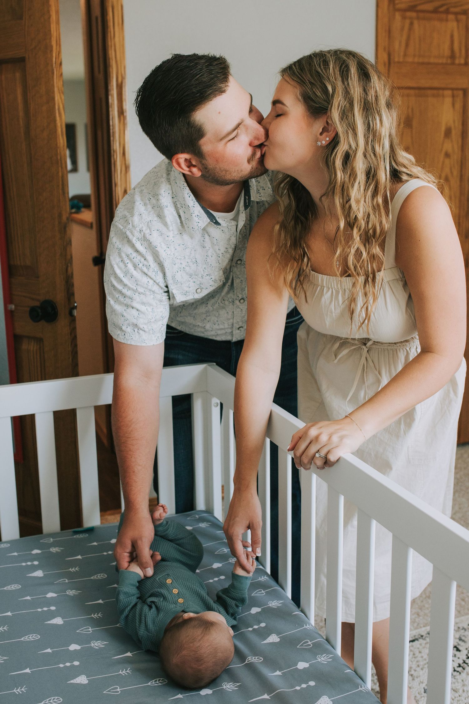 New parents sharing a kiss over their newborn baby lying in a white crib.