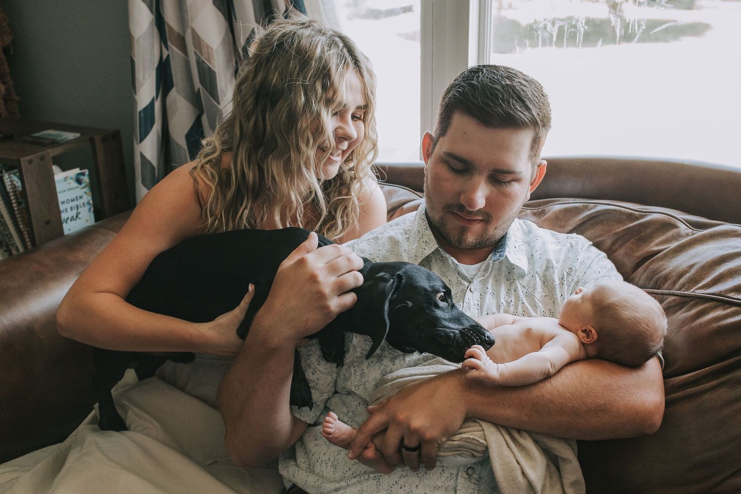 Young parents on leather couch with black dog admiring their newborn baby together.