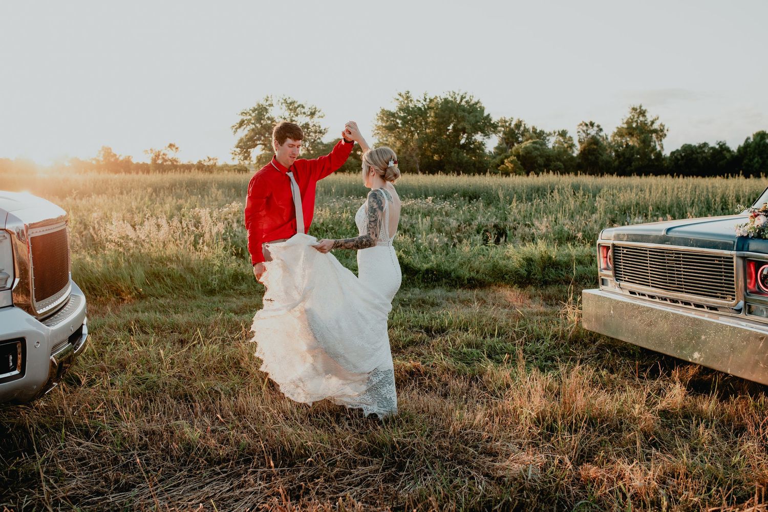 Bride and groom dancing in golden field at sunset beside vintage classic cars in rustic outdoor wedding setting.