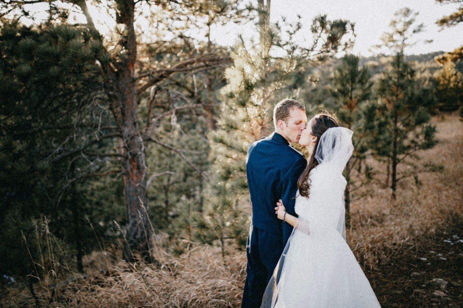 Bride and groom sharing a romantic kiss outdoors among pine trees in a scenic woodland wedding setting.