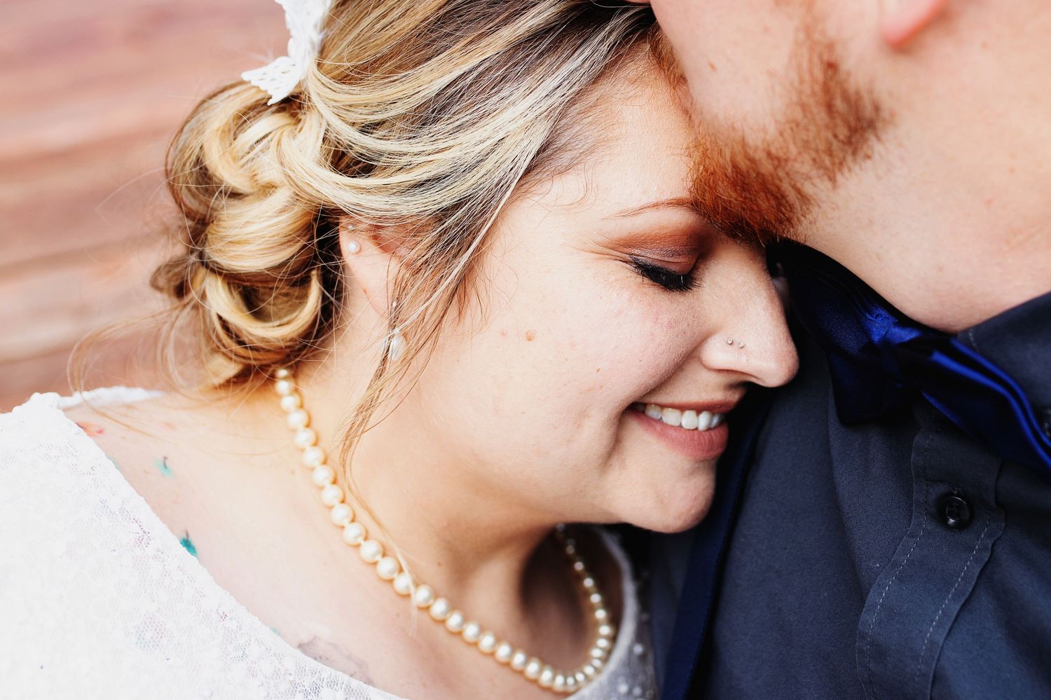 Close-up of a smiling bride wearing a pearl necklace and updo hairstyle, leaning against a groom in a navy suit.