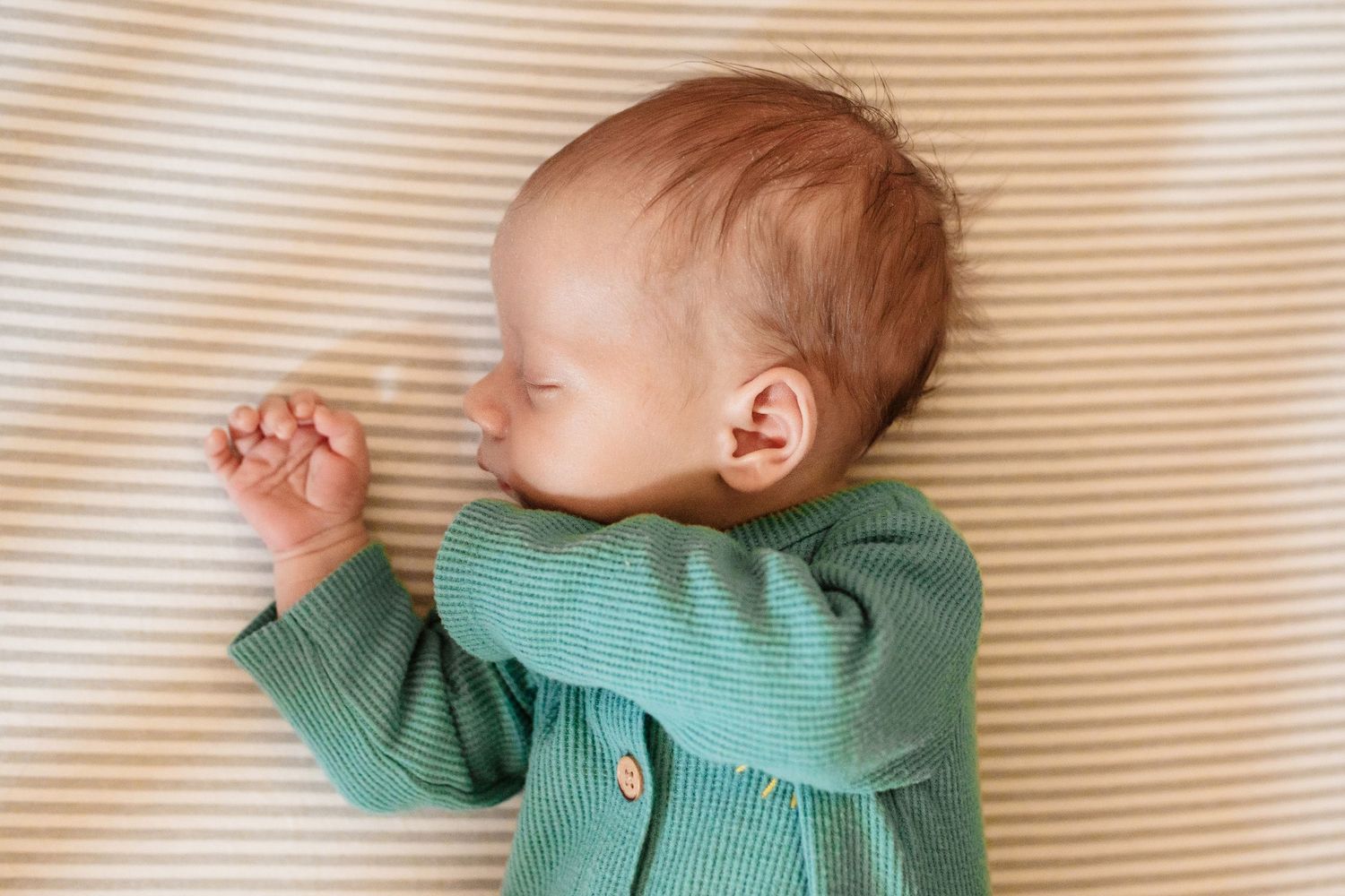 A sleeping baby wearing a mint green ribbed sweater lies on a striped cream-colored surface.