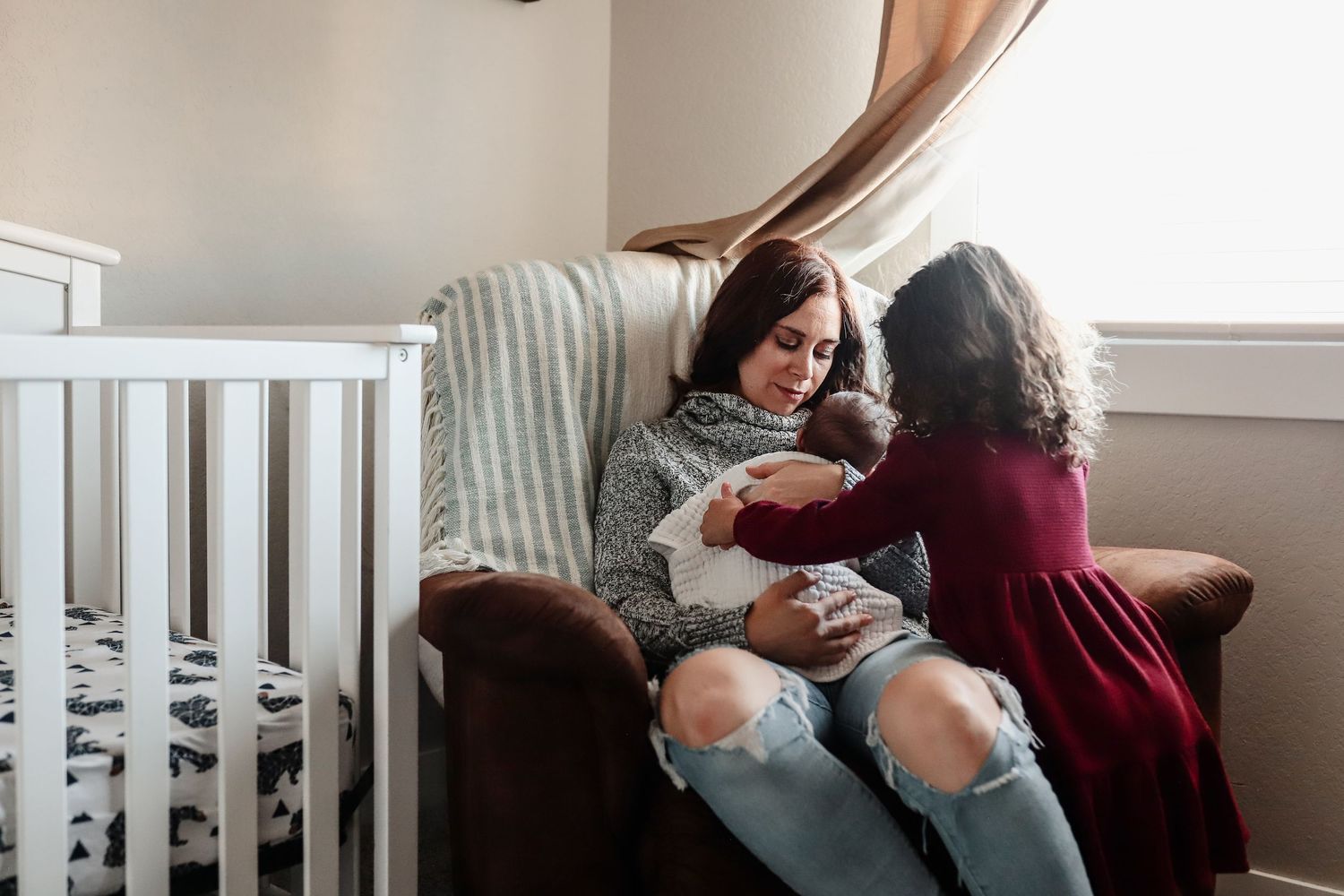 Mother sits in armchair holding newborn while young daughter in red dress reaches to touch baby.