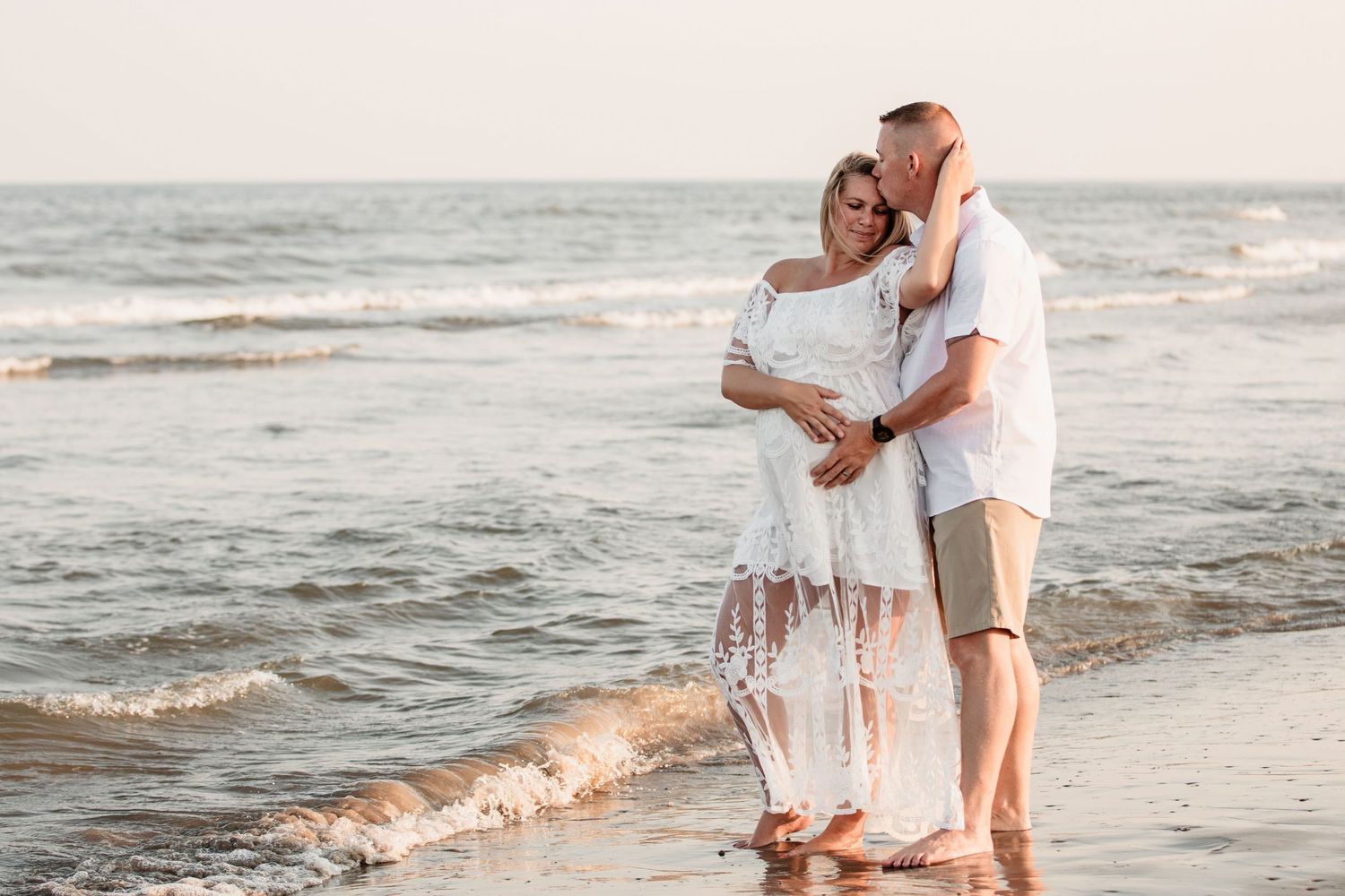 Couple sharing tender moment on beach at sunset, man kissing pregnant woman in white lace dress.